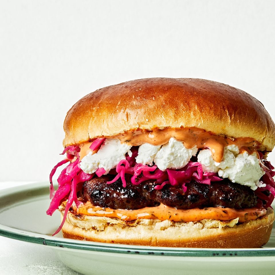 A Lamb Burger With Harissa Mayo, Pickled Red Cabbage and chèvre cheese on a brioche bun on a plate on the counter