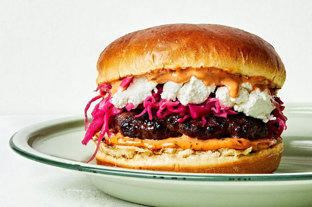 A Lamb Burger With Harissa Mayo, Pickled Red Cabbage and chèvre cheese on a brioche bun on a plate on the counter