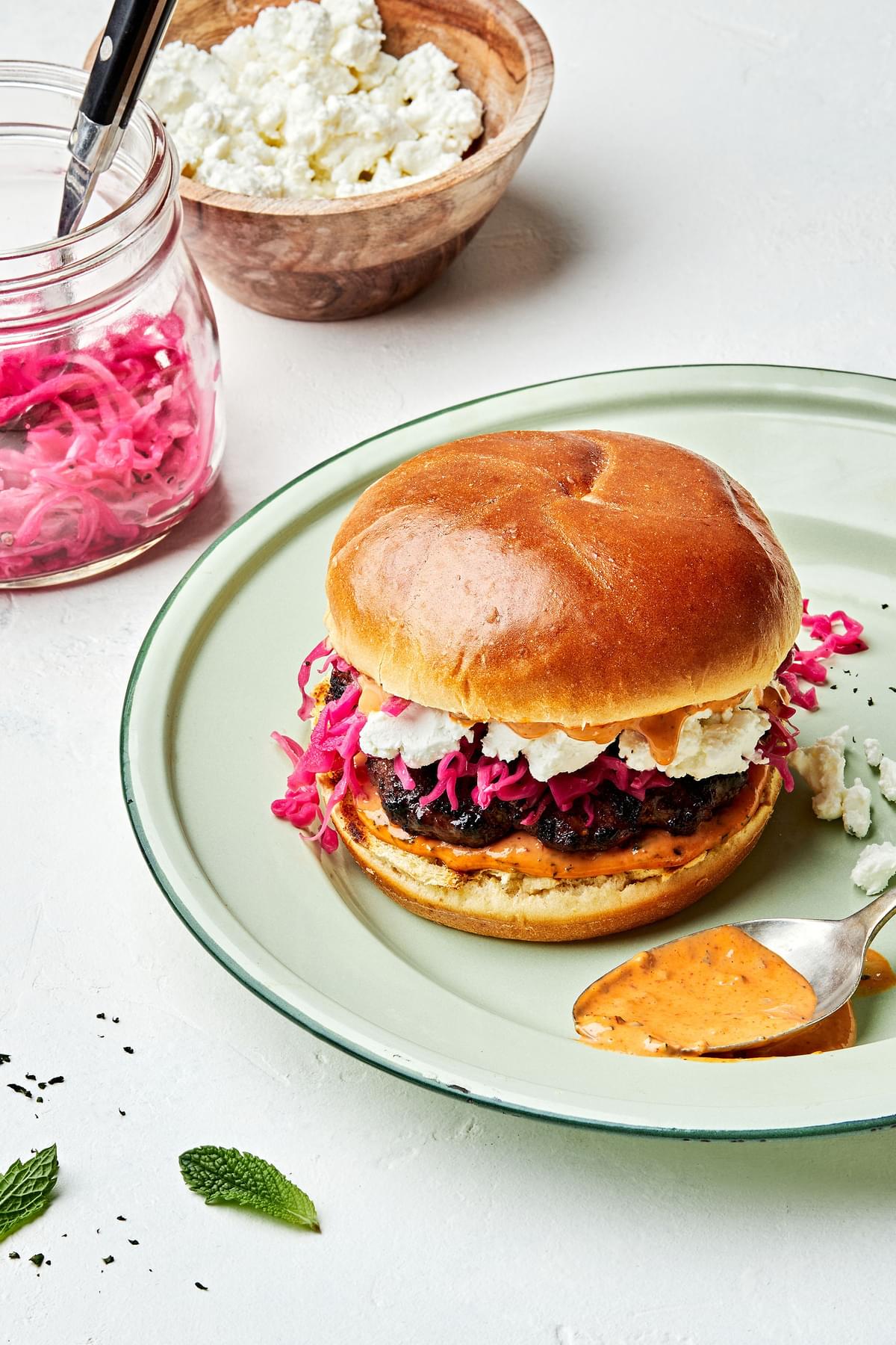 A Lamb Burger With Harissa Mayo and  Pickled Red Cabbage with chevre next to bowls of chèvre cheese and pickled red cabbage