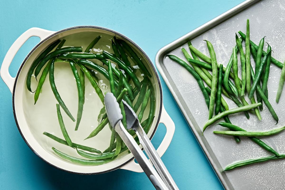 blanched green beans in a pot being transferred to a baking sheet with tongs to finish cooking in the oven