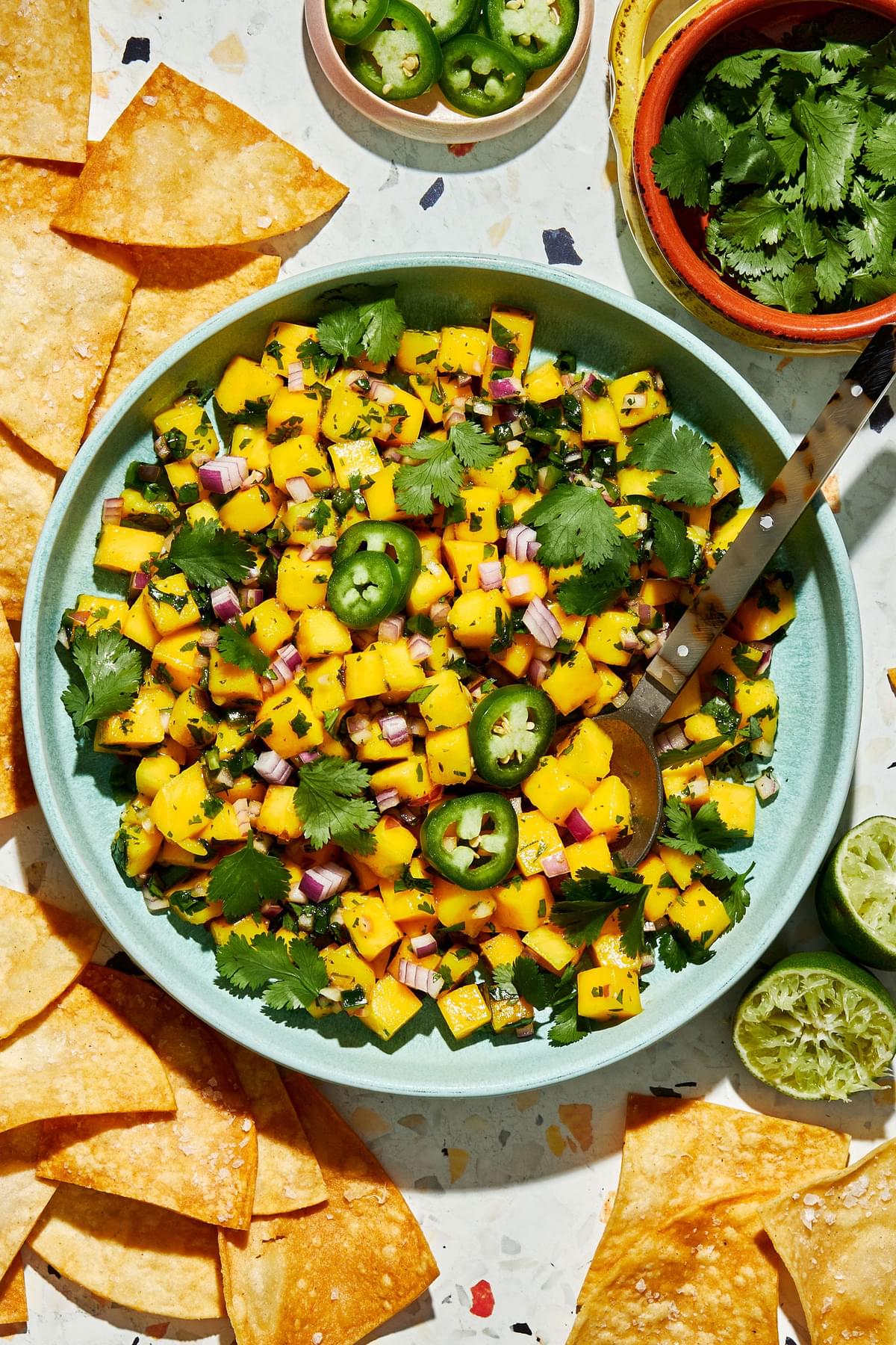 mango salsa in a serving bowl with a spoon surrounded by chips and bowls of chopped cilantro and jalapeño