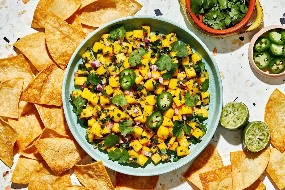 mango salsa in a serving bowl with a spoon surrounded by chips and bowls of chopped cilantro and jalapeño