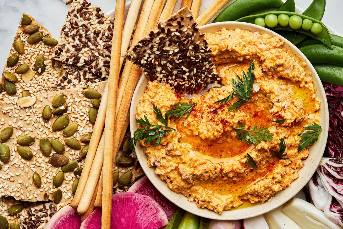 homemade nutty carrot dip in a bowl on top of a platter surrounded with raw peas, cucumbers, radishes and crackers