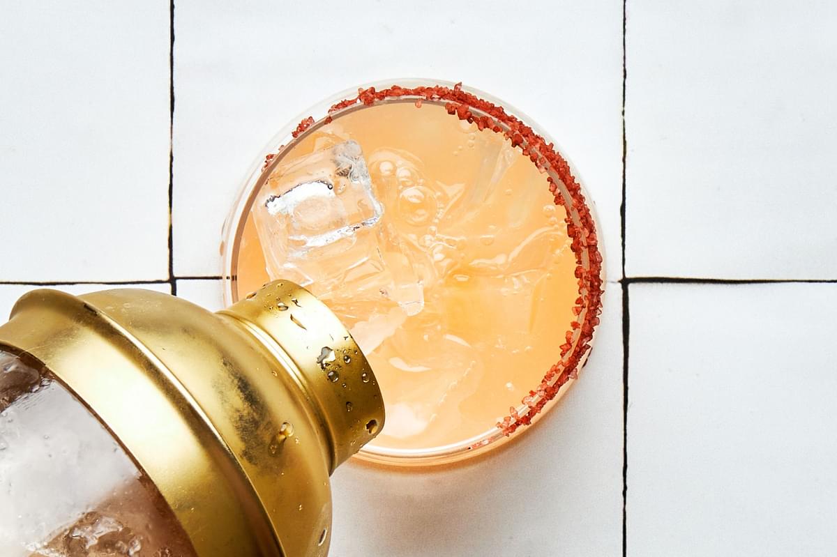 A Paloma being poured from a cocktail shaker into a salt lined glass full of ice made with tequila, grapefruit & lime juices