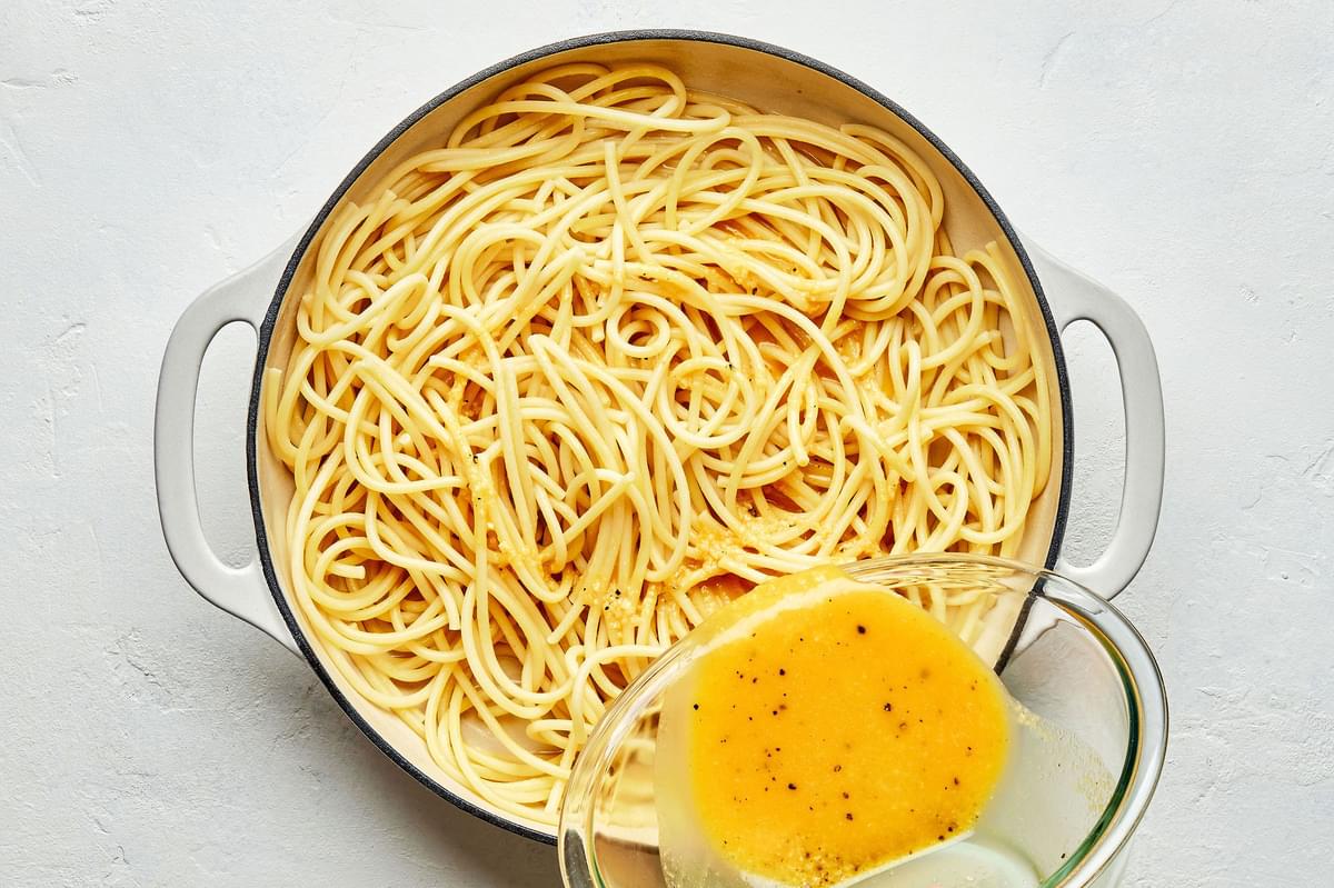 whisked eggs, Romano and pepper being slowly poured over cooked spaghetti noodles with rendered fat in a skillet