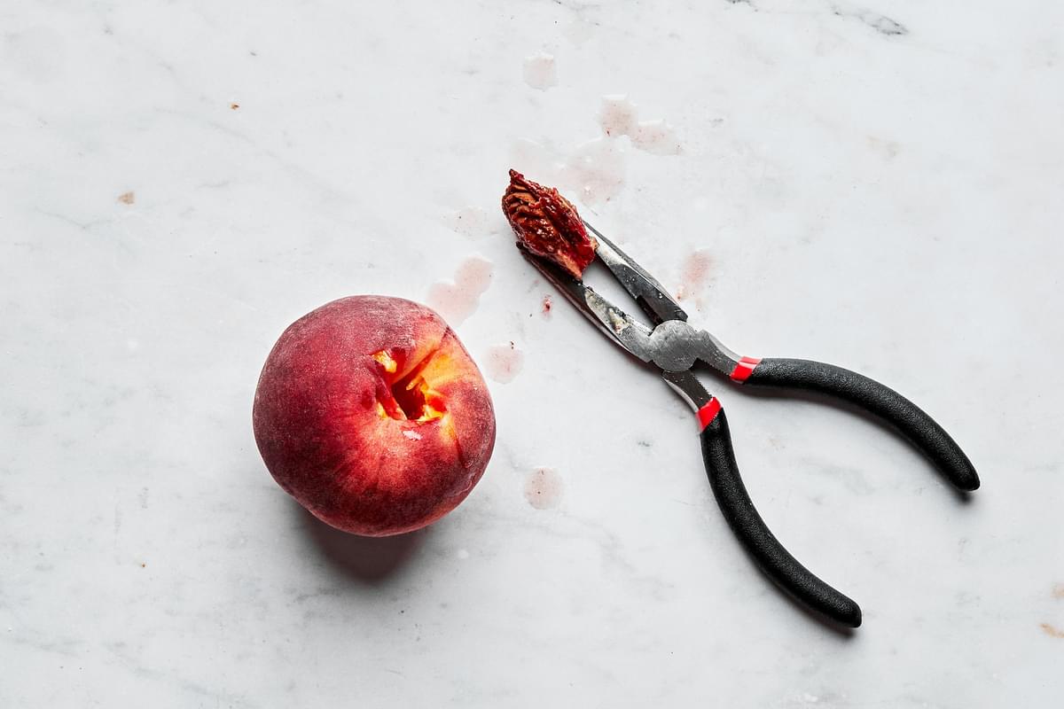 a peach with the pit removed next to a pair of needle nosed pliers holding the pit