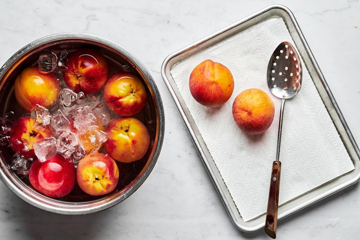blanched peaches in an ice bath next to a paper towel lined baking sheet with a couple of peaches and a slotted spoon