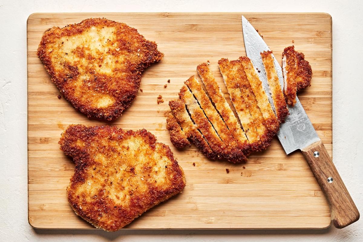 pan-fried breaded pork chops being sliced with a knife on a cutting board to use for pork milanese bowls