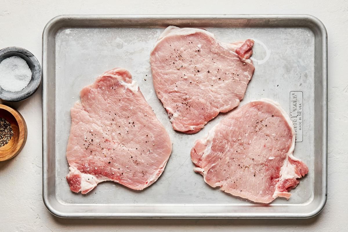 raw pounded boneless pork chops on a baking sheet seasoned with salt and pepper