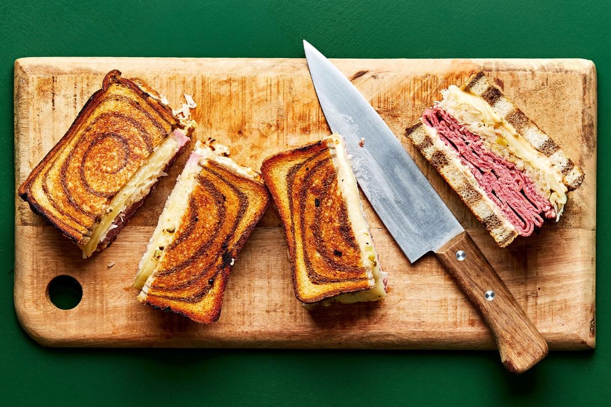 2 reuben made with rye bread, pastrami, sauerkraut, Swiss cheese and dressing being cut in half with a knife on a bread board