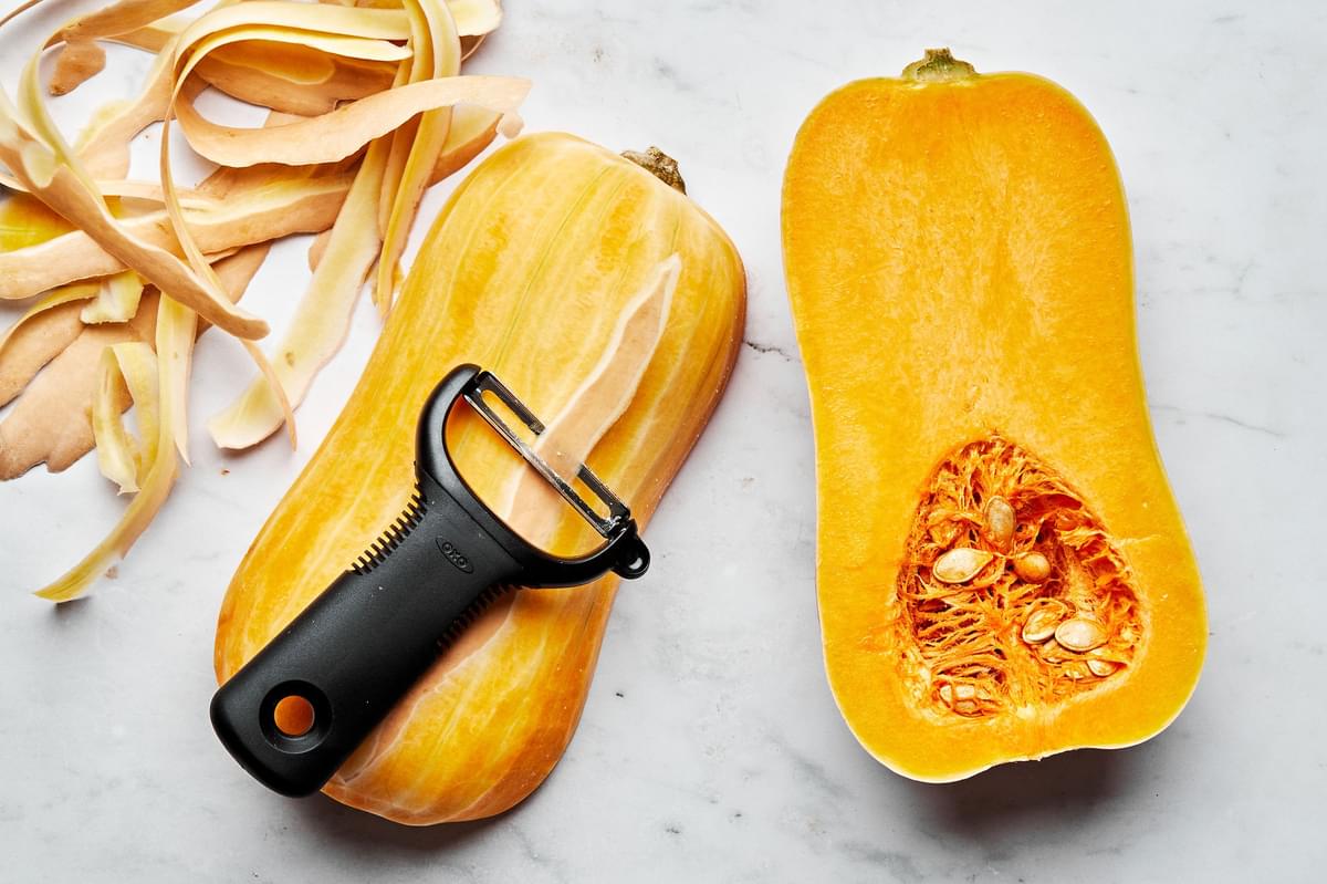 a raw butternut squash cut in half lengthwise on the counter being peeled using a vegetable peeler