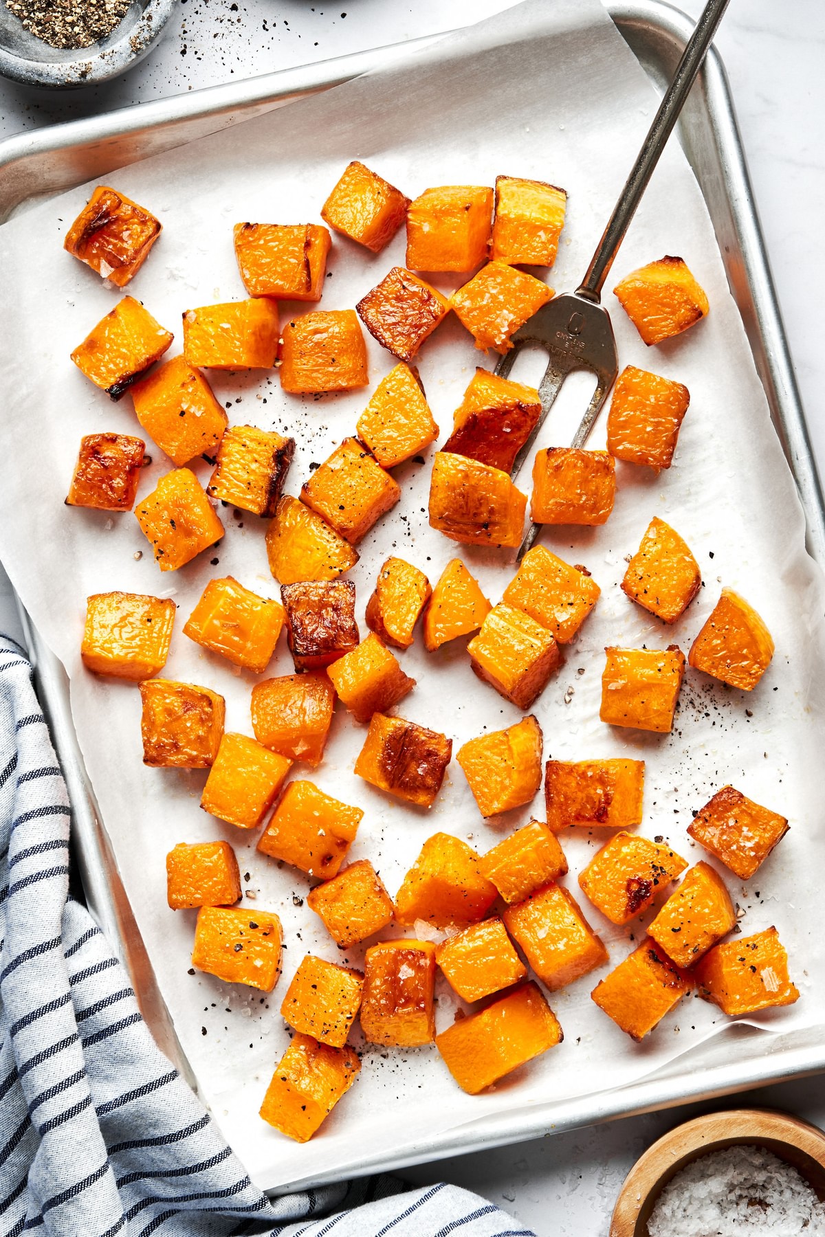 cubed roasted butternut squash seasoned with olive oil, salt and pepper on a baking sheet with a serving fork