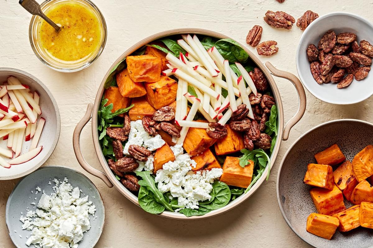 A Roasted Sweet Potato Salad surrounded by bowls of apples, goat cheese, candied pecans and orange vinaigrette