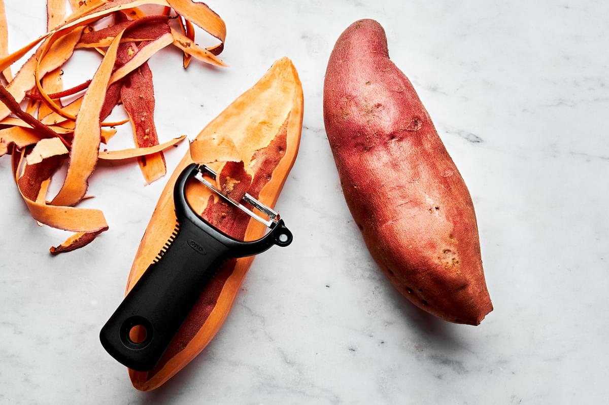 raw sweet potatoes being peeled with a vegetable peeler