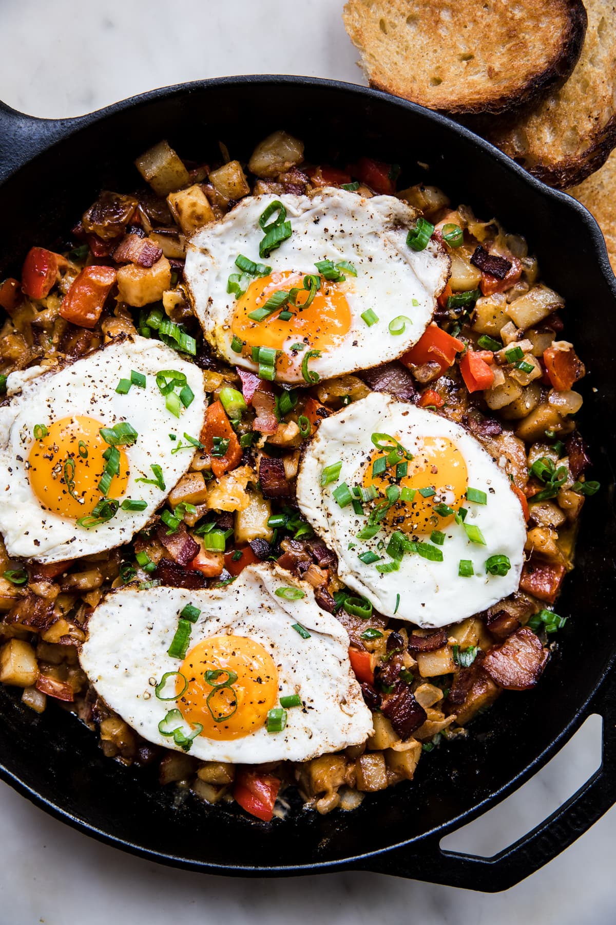 Black skillet with bacon, potato and cheddar hash topped with fried eggs and green onions.