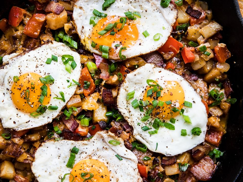 Black skillet with bacon, potato and cheddar hash topped with fried eggs and green onions.