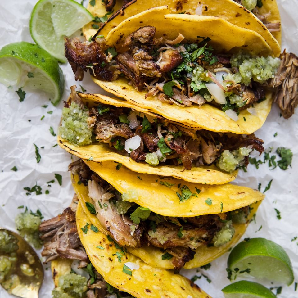 five carnitas tacos on white parchment paper topped with lime juice, cilantro and onions