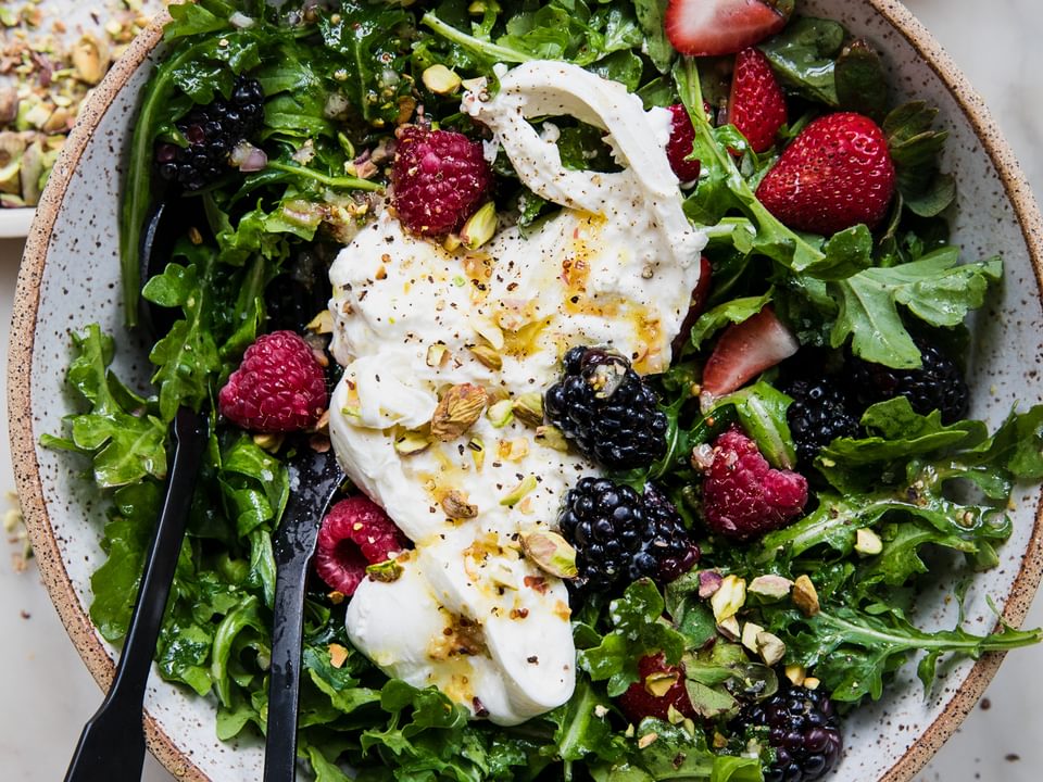 Large bowl of arugula dressing with honey mustard dressing and topped with pistachios, burrata and salt and pepper
