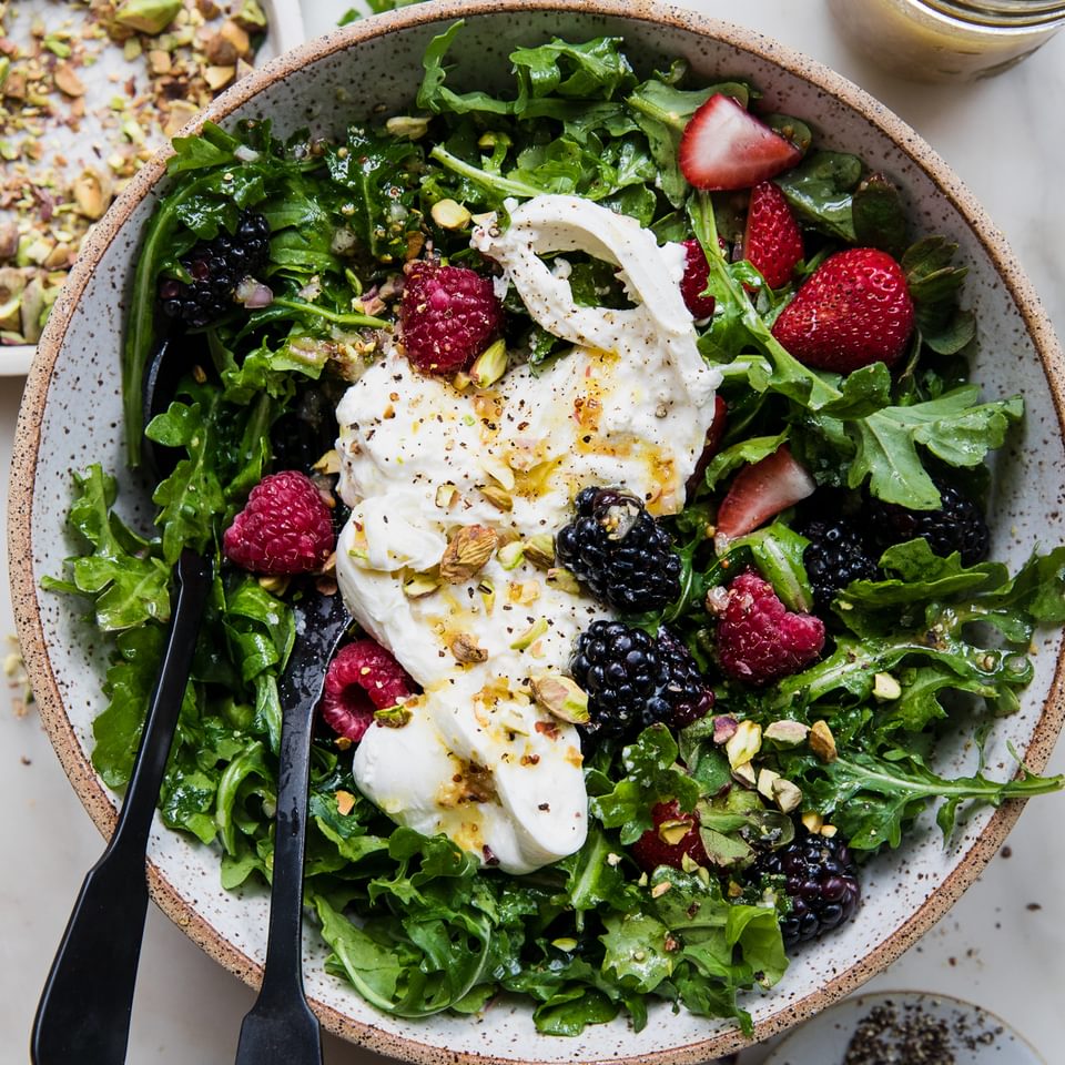 Large bowl of arugula dressing with honey mustard dressing and topped with pistachios, burrata and salt and pepper