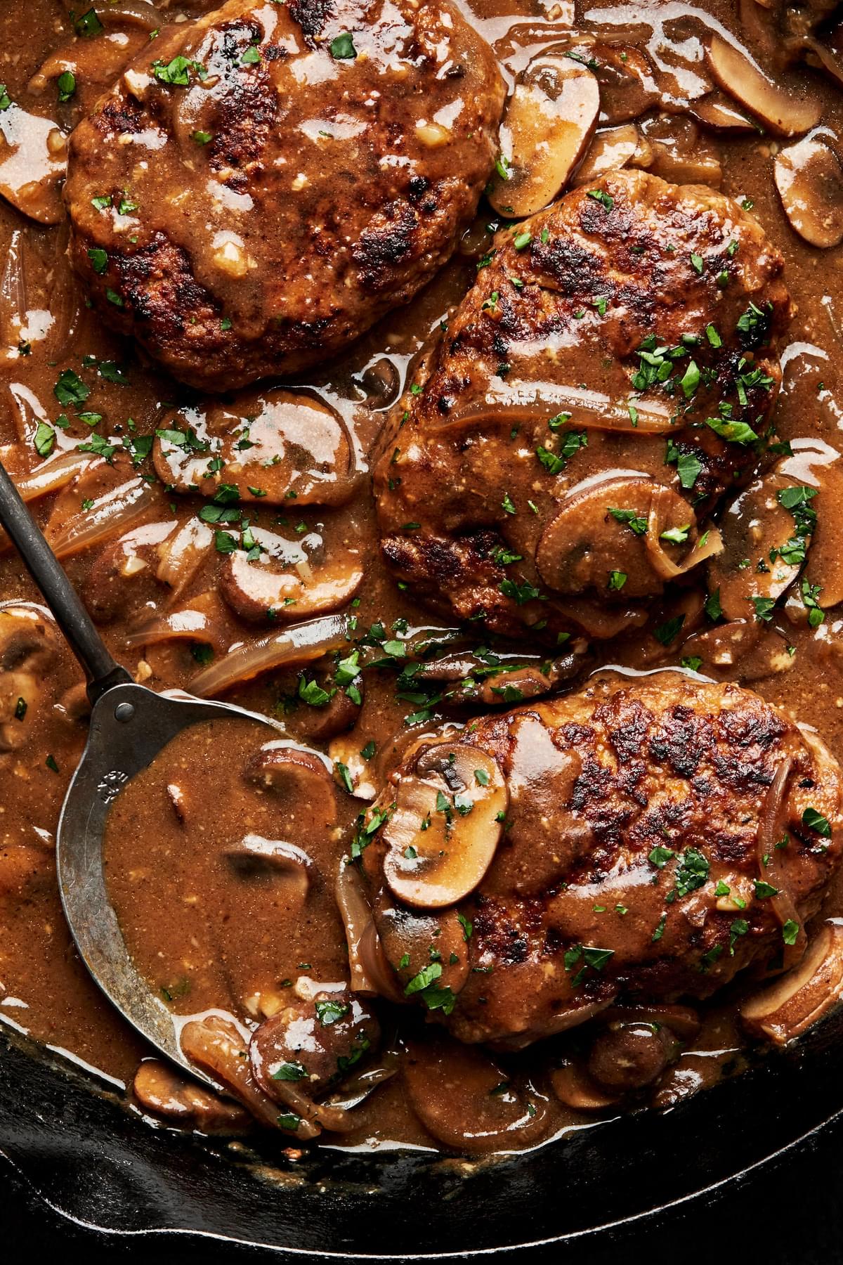 a cast iron skillet filled with salisbury steaks in a homemade gravy with mushrooms and onions topped with fresh parsley