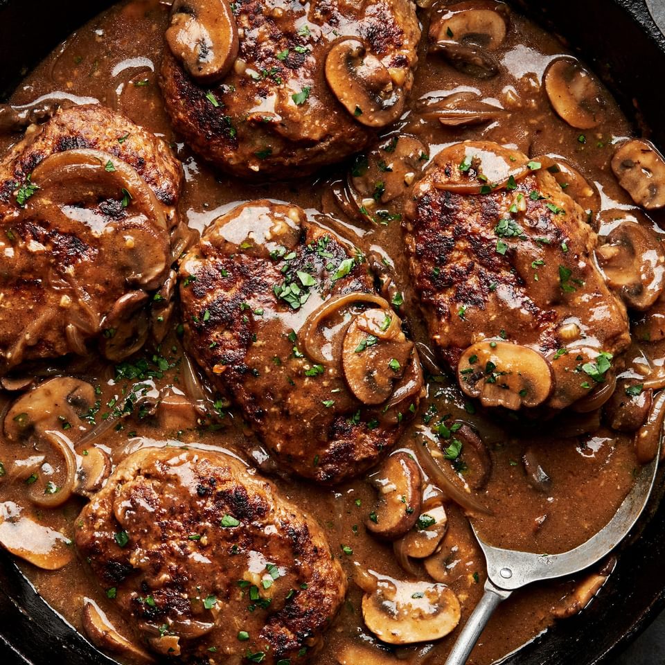 5 salisbury steaks in a homemade gravy with mushrooms and onions topped with fresh parsley in a cast iron skillet with spoon