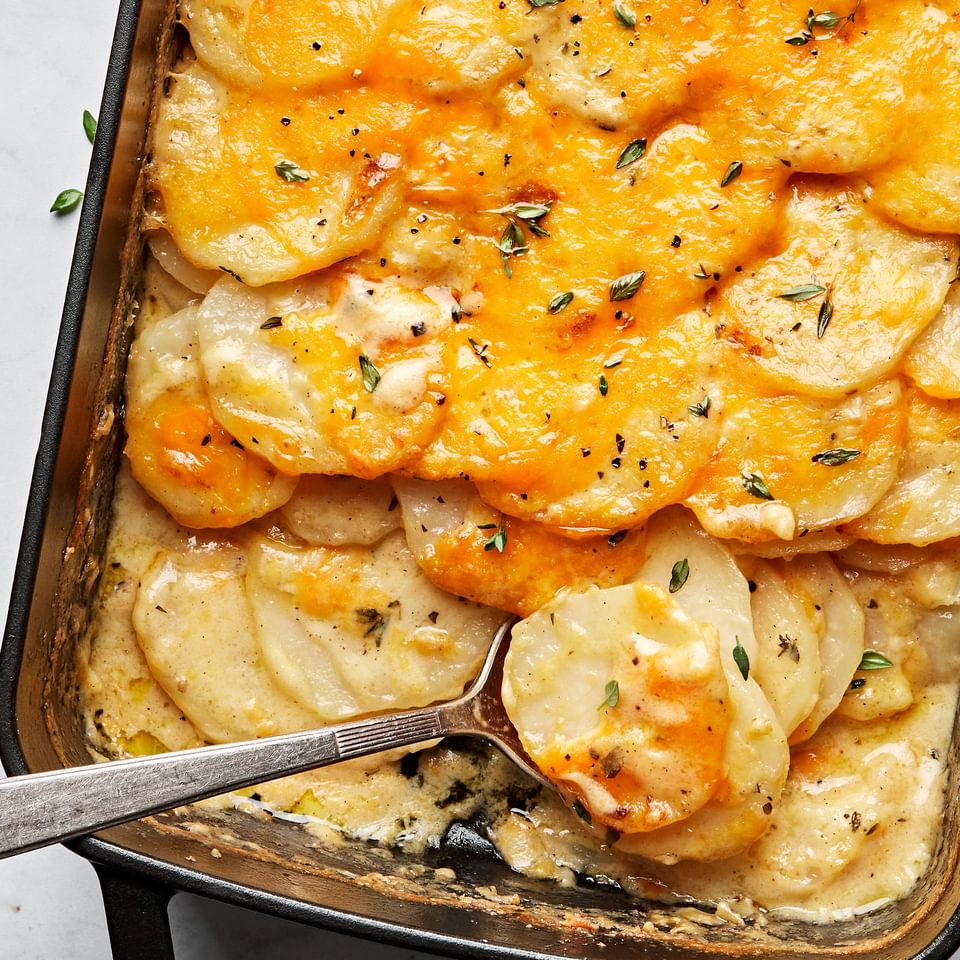 homemade scalloped potatoes covered in melted cheese in a casserole dish being scooped out with a spoon