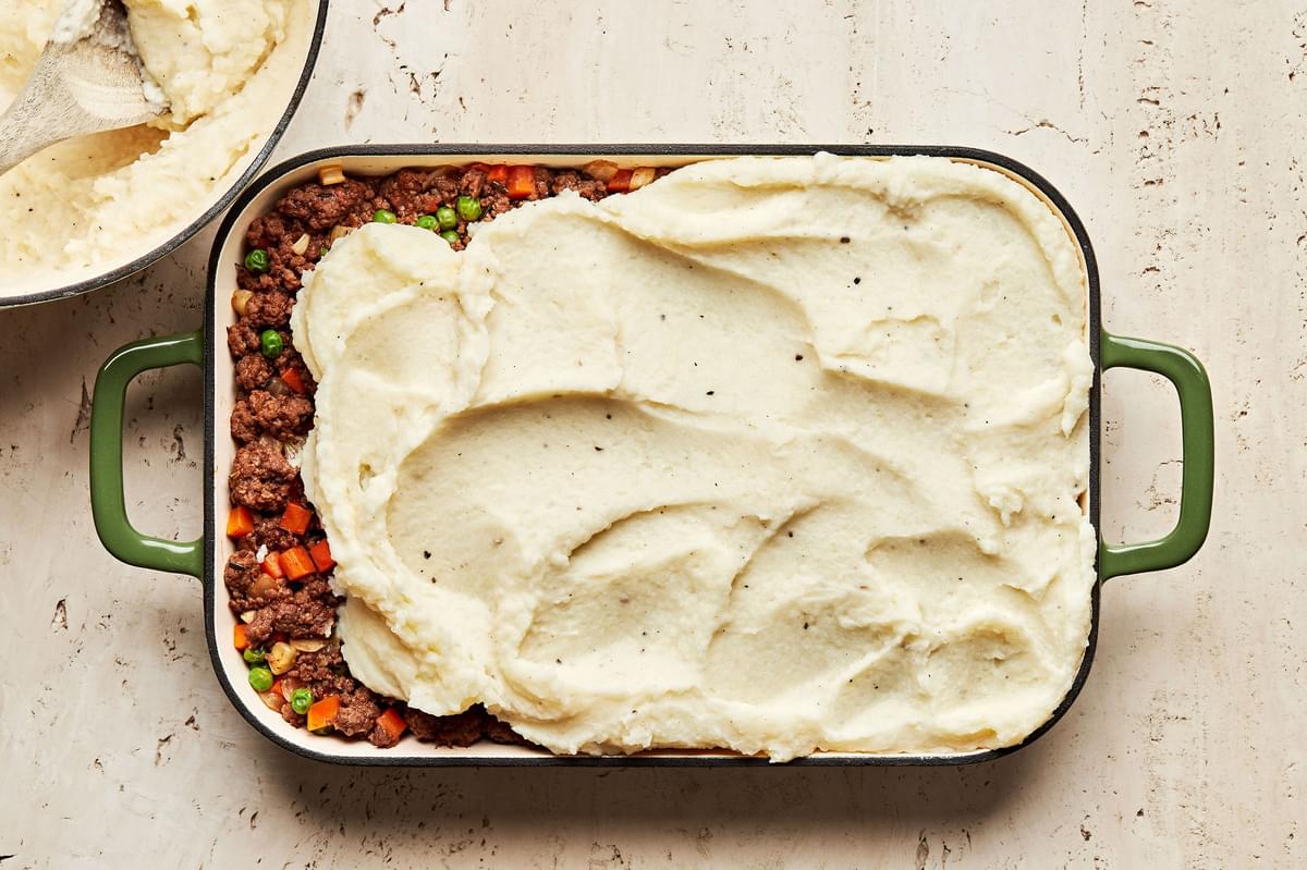 mashed potatoes being spread on top of a ground beef and vegetable mixture for in a casserole dish to make shepherd's pie