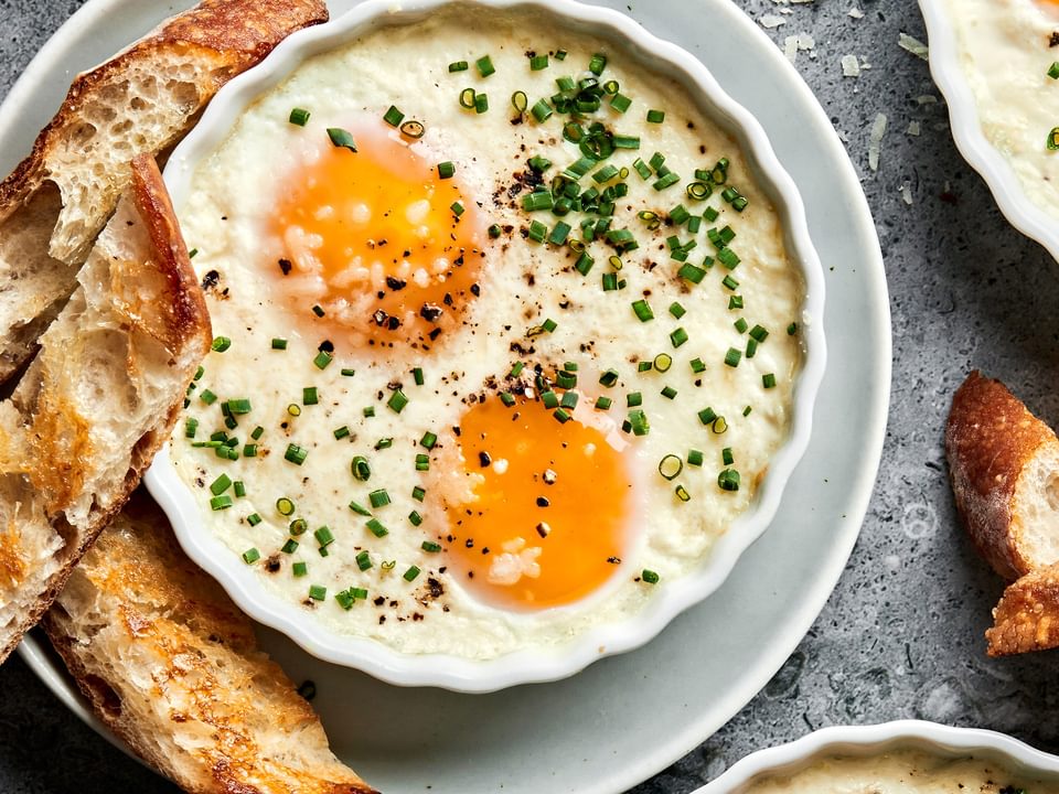 ramekins of shirred eggs garnished with chopped chives and served with crusty bread next to a bowl of parmesan