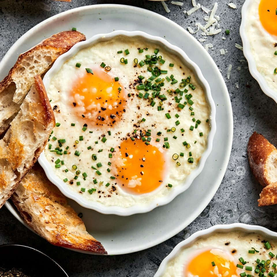 ramekins of shirred eggs garnished with chopped chives and served with crusty bread next to a bowl of parmesan