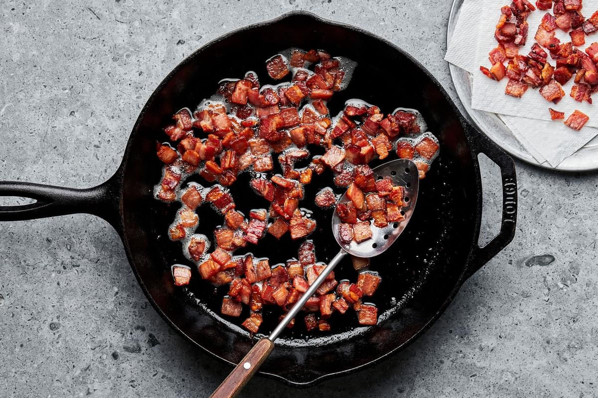 cooked diced bacon being spooned out of a skillet with a slotted spoon