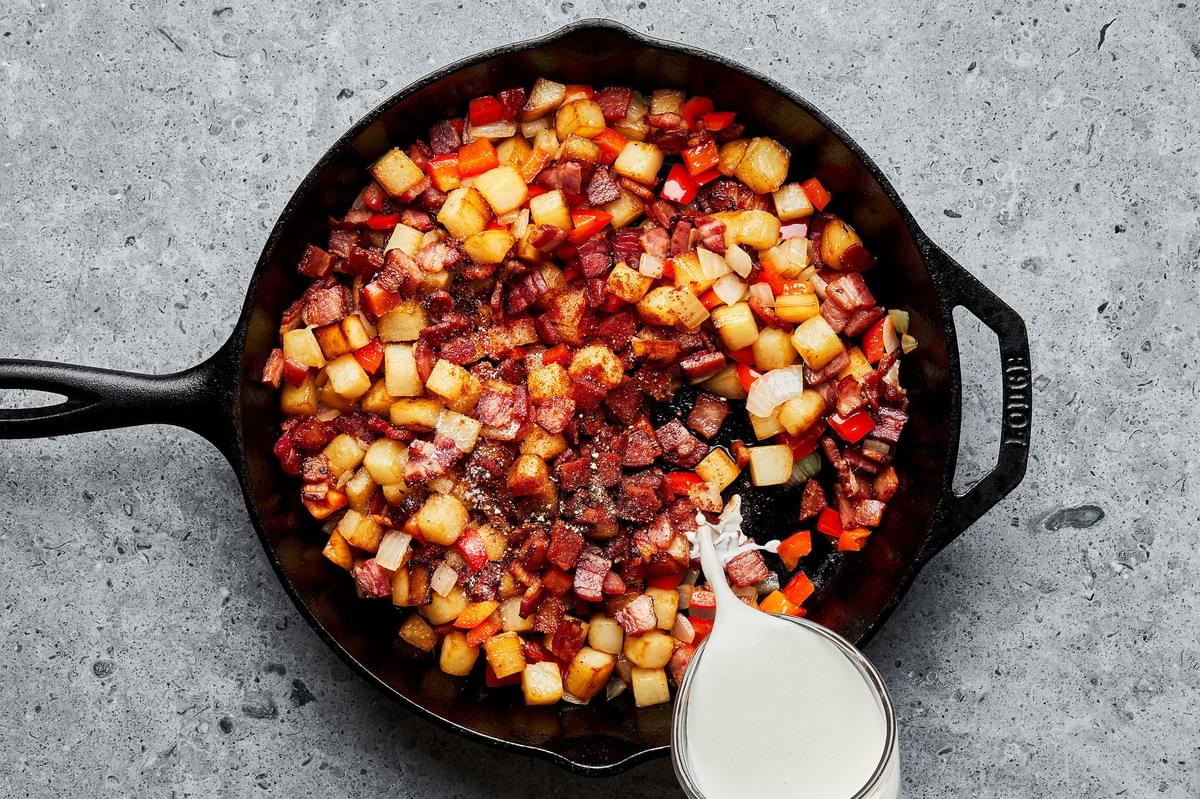 cream being poured into a skillet with bacon, potatoes, onion, bell pepper, worcestershire sauce, paprika, salt and pepper