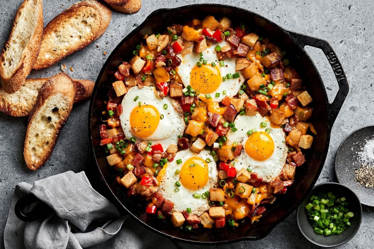Smoky bacon cheddar breakfast hash topped with fried eggs in a skillet being served along side crusty bread