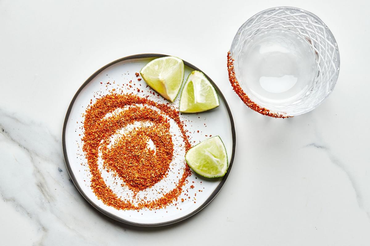 the rim of a rocks glass dipped in Tajín seasoning next to a plate of Tajín seasoning and lime wedges for a spicy margarita