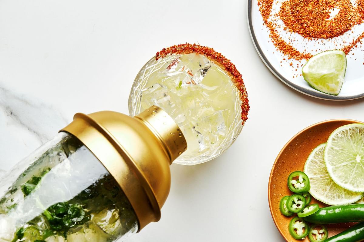 a spicy margarita in a cocktail shaker being poured into a rocks glass with a tajín seasoning rim