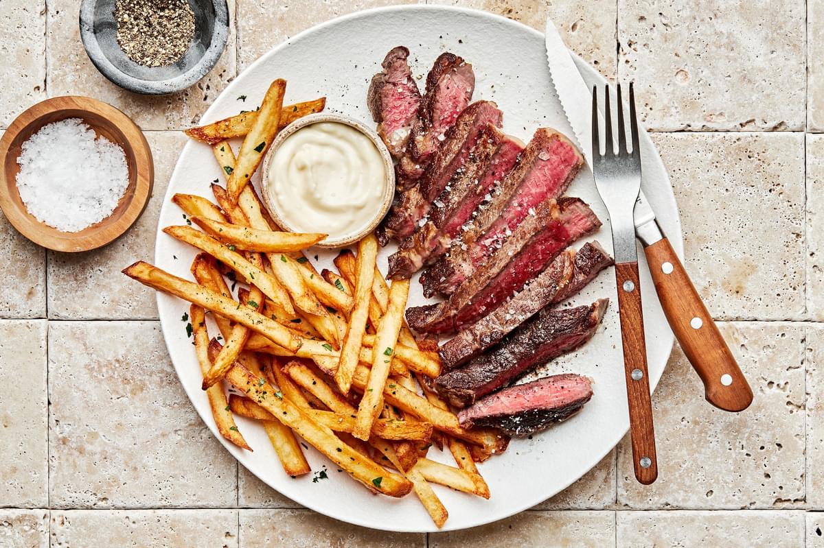 Homemade Steak Frites on plate with a fork and knife served with garlic aioli next to bowls of salt and pepper for seasoning
