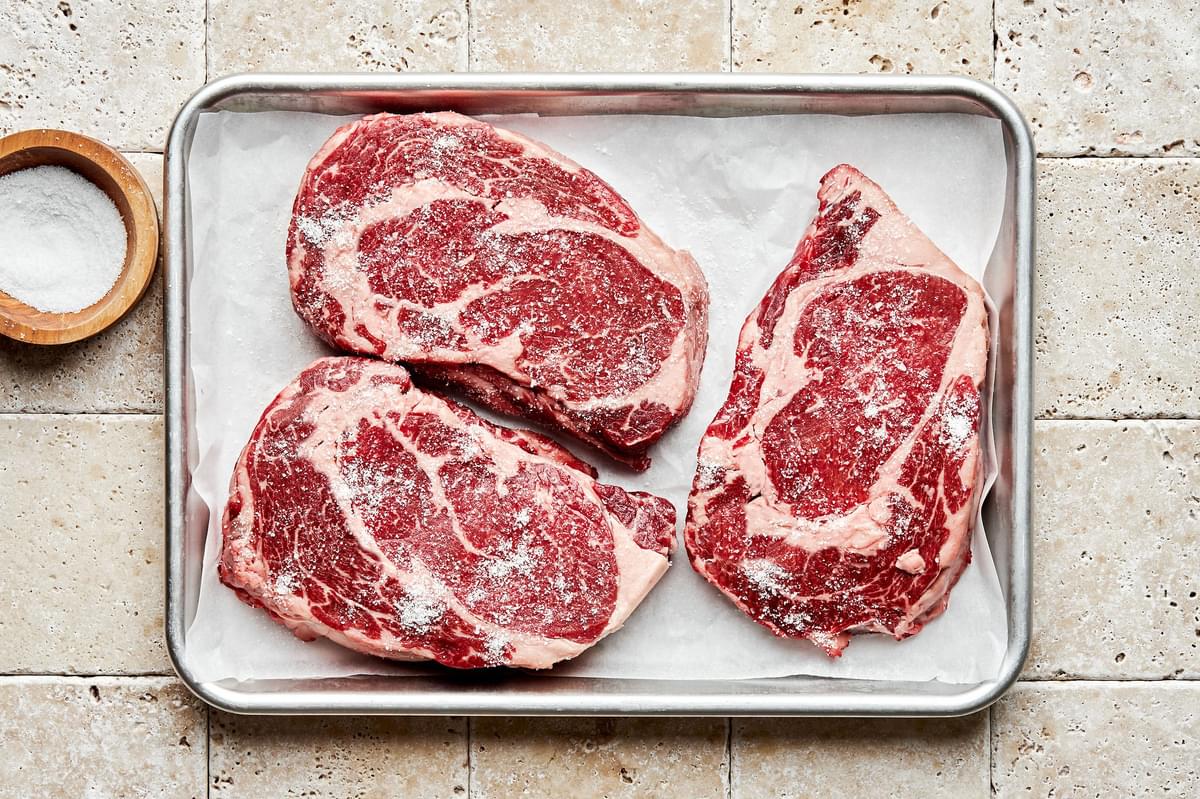 raw rib eye steaks patted dry and seasoned with salt on a baking sheet next to a bowl of salt