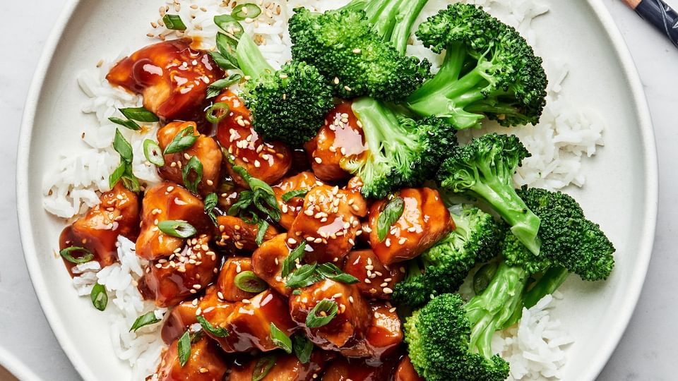 homemade teriyaki chicken on a plate served over white rice with steamed broccoli on the side next to chop sticks