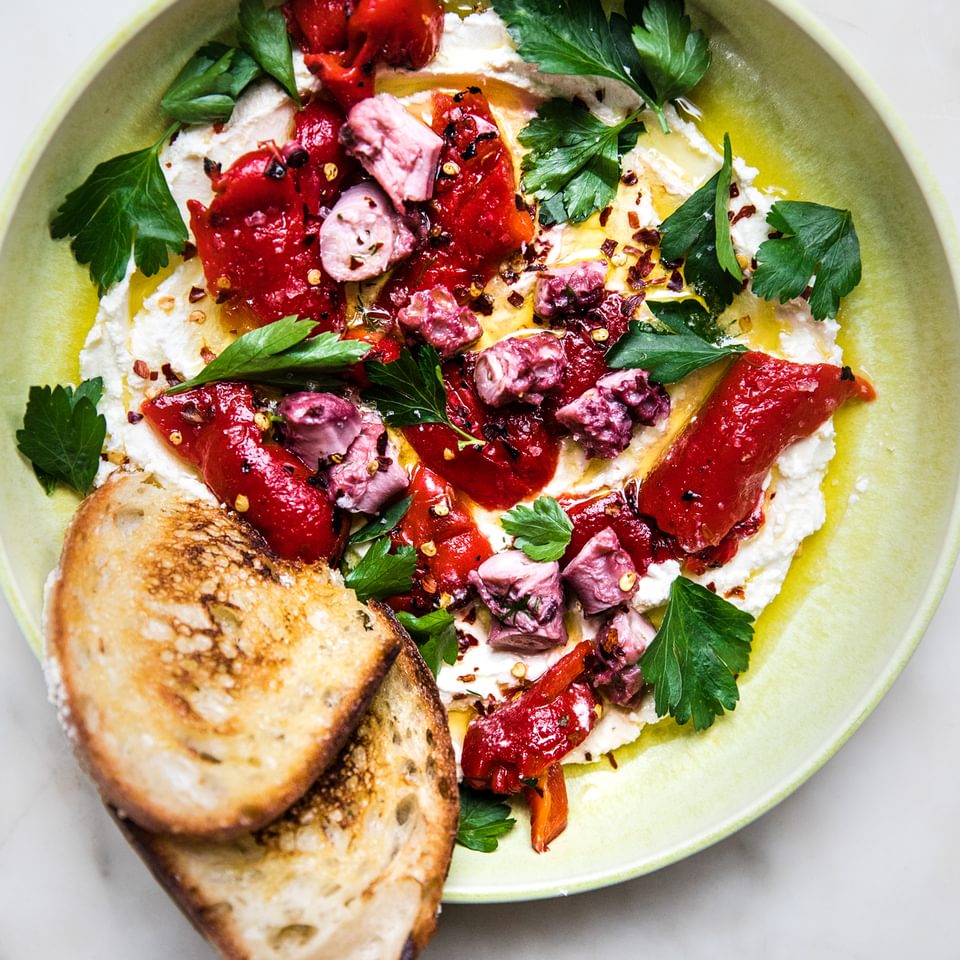 Yellow bowl with whipped feta, roasted red pepper and parsley topped with octopus served with toast.