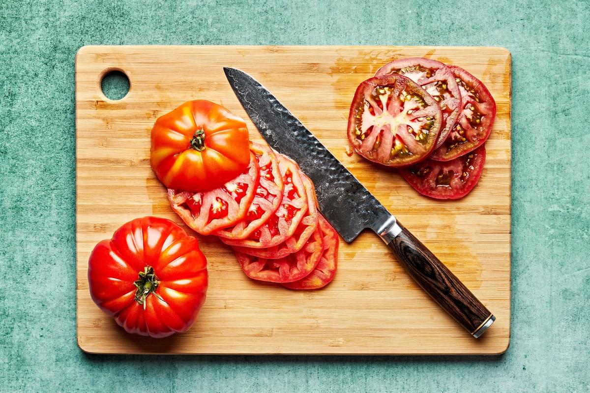 heirloom tomatoes being thinly sliced on a cutting board with a sharp knife