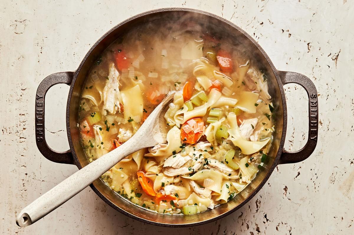a pot of homemade turkey soup being stirred with a wooden spoon made with turkey, noodles, vegetables and spices