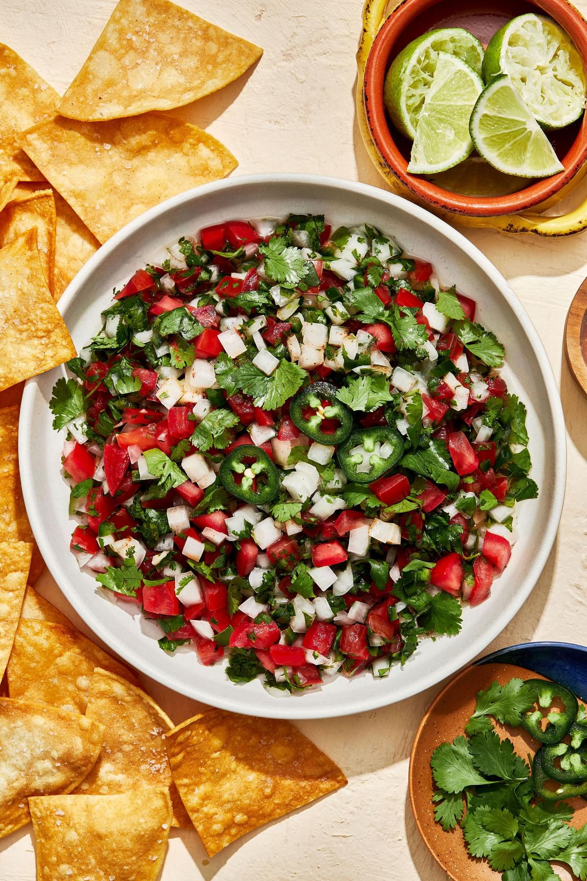 homemade pico de Gallo in a serving bowl surrounded by tortilla chips for serving and bowls of lime wedges and cilantro