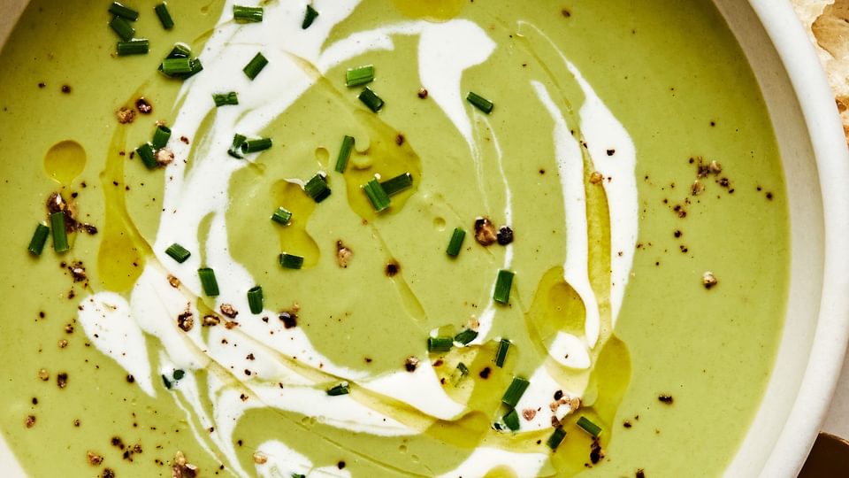 a bowl of homemade cream of broccoli soup sprinkled with chives and garnished with a swirl of cream served with crusty bread