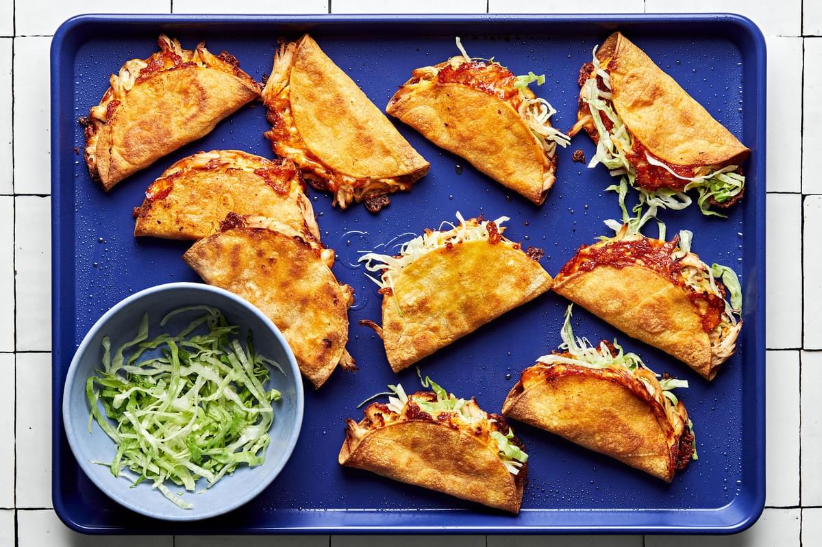 10 warm crispy chicken tacos on a baking sheet next to a bowl of shredded lettuce for topping