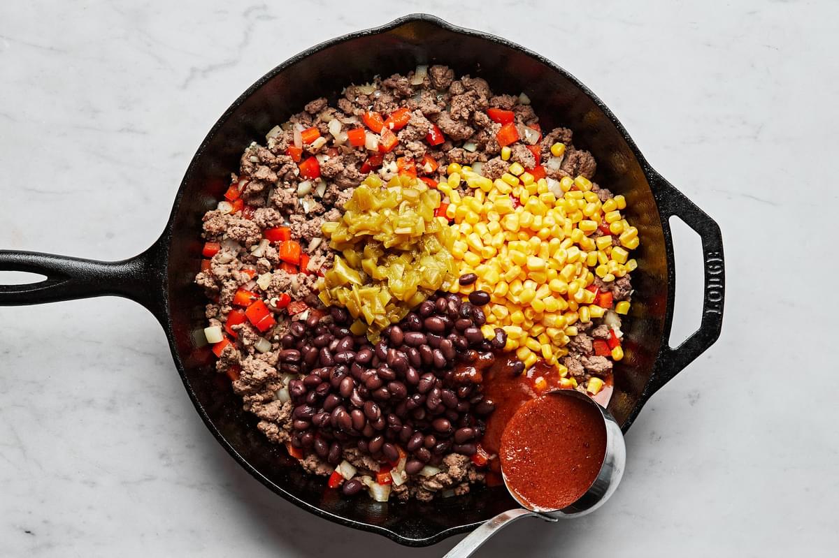 enchilada sauce being poured over ground beef, onion, peppers, chiles, corn and black beans in a skillet