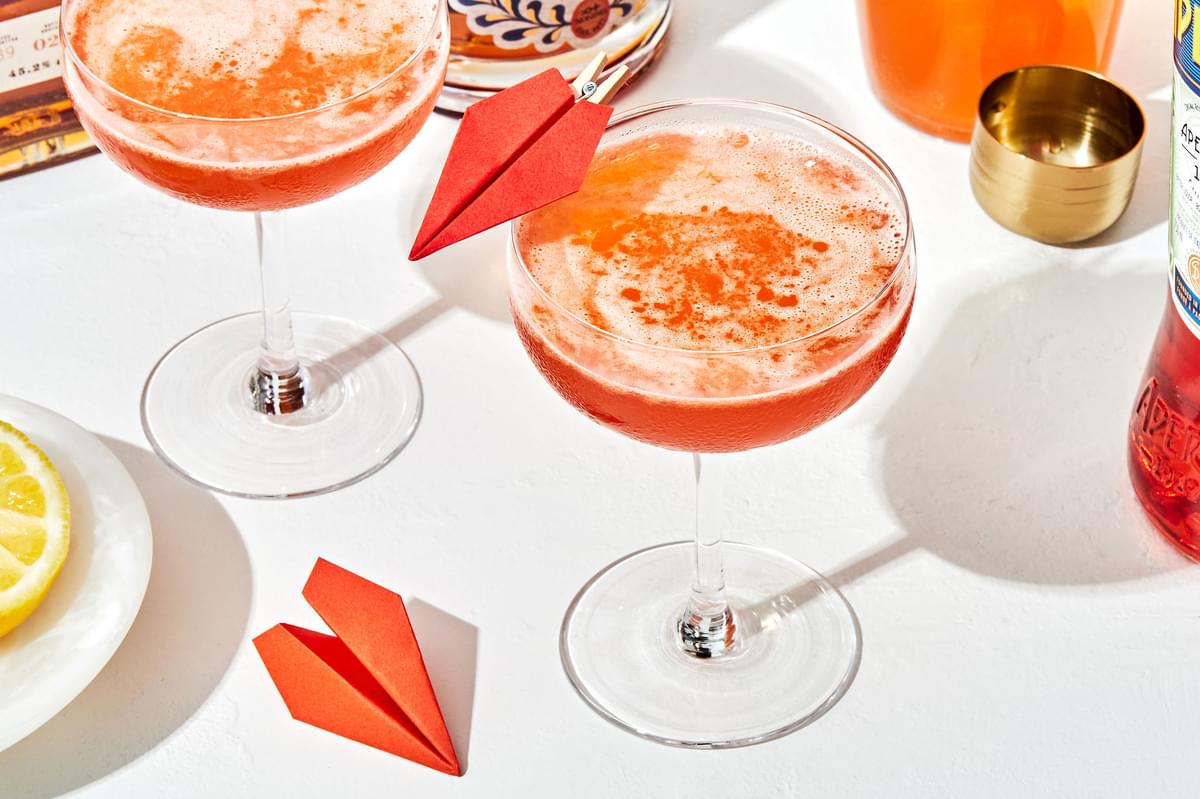 a paper plane cocktail in a coupe glass with a small paper airplane attached to the side of the glass