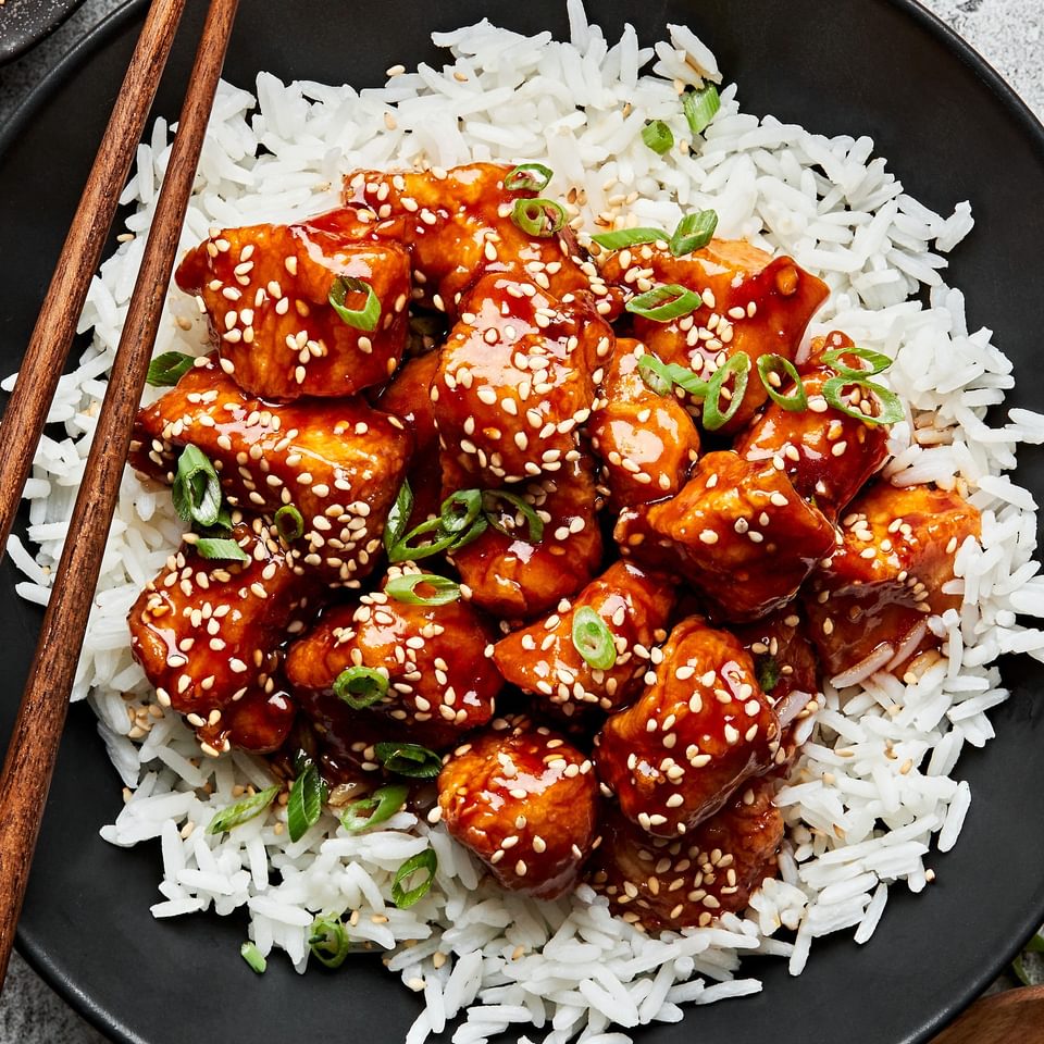 a bowl of homemade sesame chicken on top of white rice surrounded by bowl of green onions, sesame seeds for garnishing