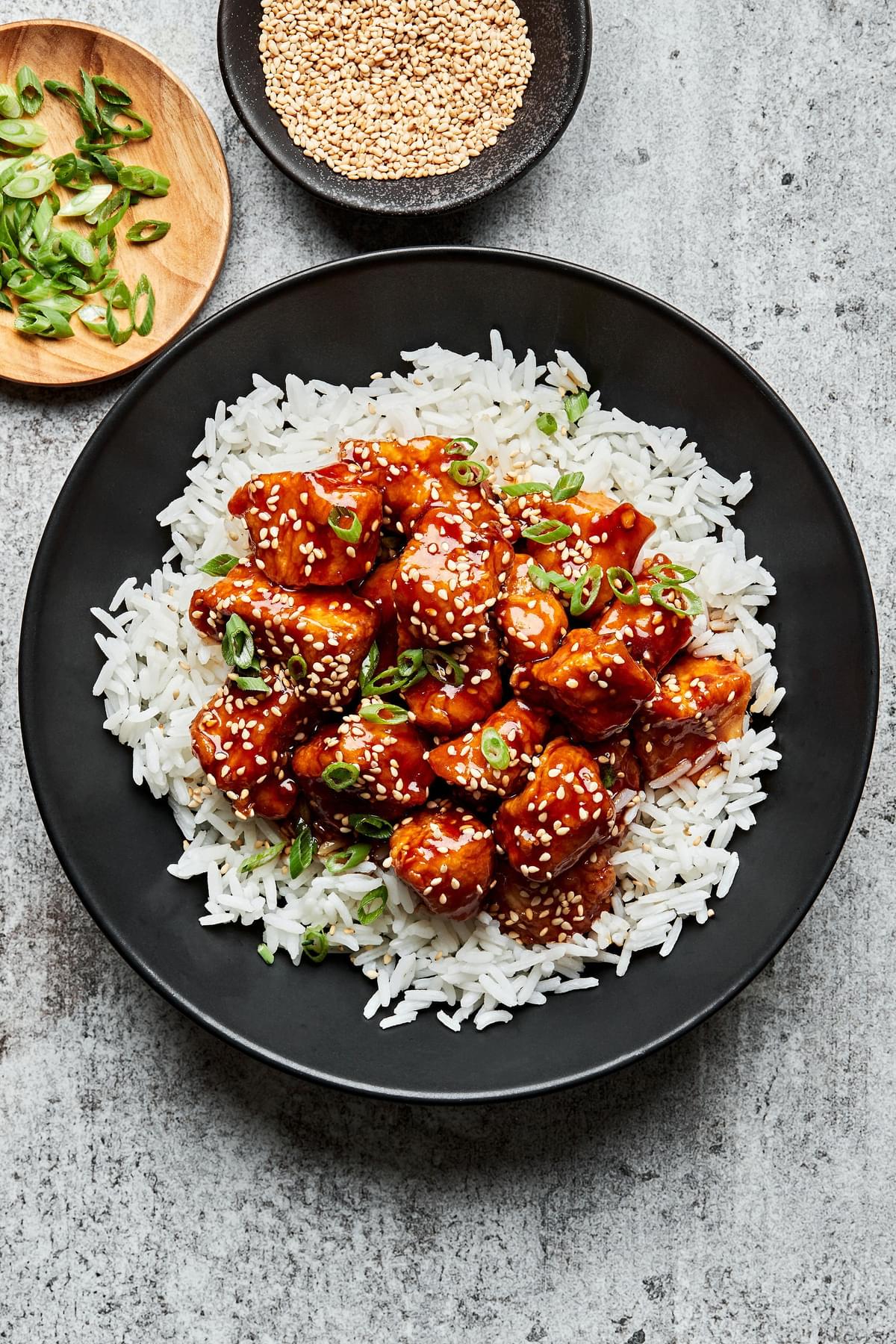 a bowl of homemade sesame chicken on top of white rice surrounded by bowl of green onions, sesame seeds for garnishing