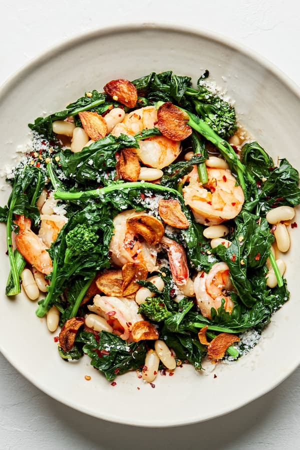 spicy broccoli rabe with white beans and shrimp in a bowl served with crusty bread topped with parmesan and fried garlic