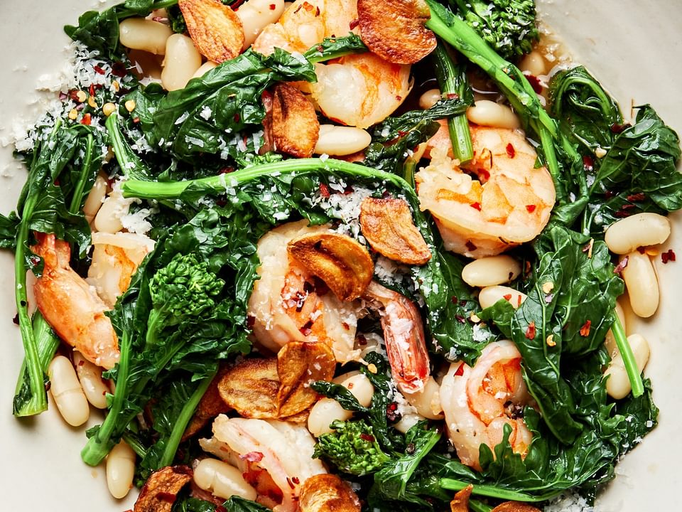 spicy broccoli rabe with white beans and shrimp in a bowl served with crusty bread topped with parmesan and fried garlic