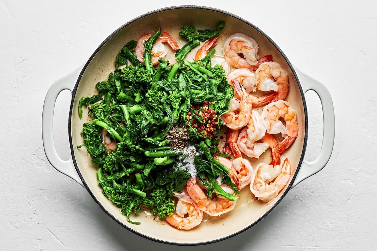 cooked shrimp and blanched broccoli rabe cooking in a skillet seasoned with salt, pepper and crushed red pepper flakes